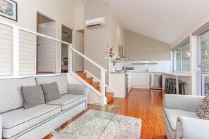 A modern living room with a grey sofa, a glass coffee table, and a staircase leading to a kitchen area. - Wine Country Villas (Pokolbin)
