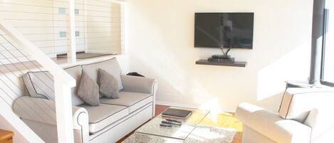 A modern living room with a staircase, a sofa, a glass coffee table, and a flat-screen TV mounted on the wall.