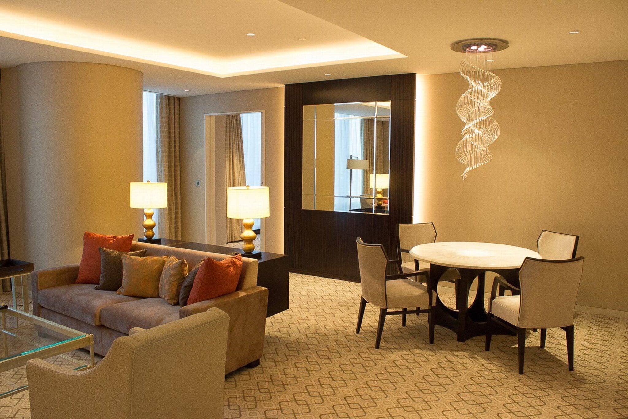 A hotel room with a sofa, a dining table with chairs, a mirror, and a patterned carpet.