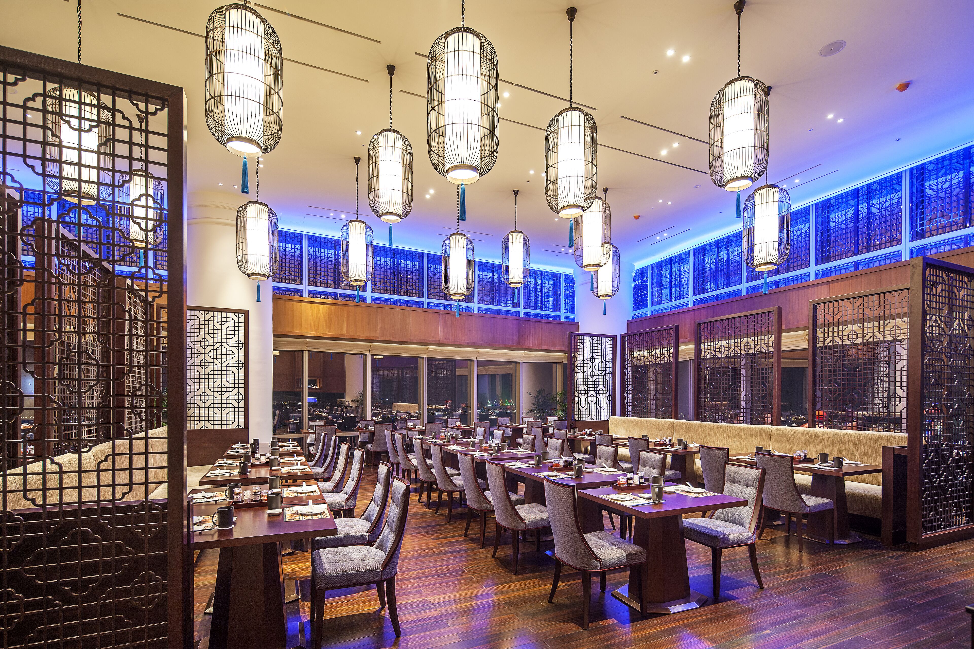 A spacious restaurant with multiple dining tables and chairs, hanging lantern-style lights, and large windows with decorative screens.