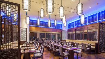 A spacious restaurant with multiple dining tables and chairs, hanging lantern-style lights, and large windows with decorative screens.