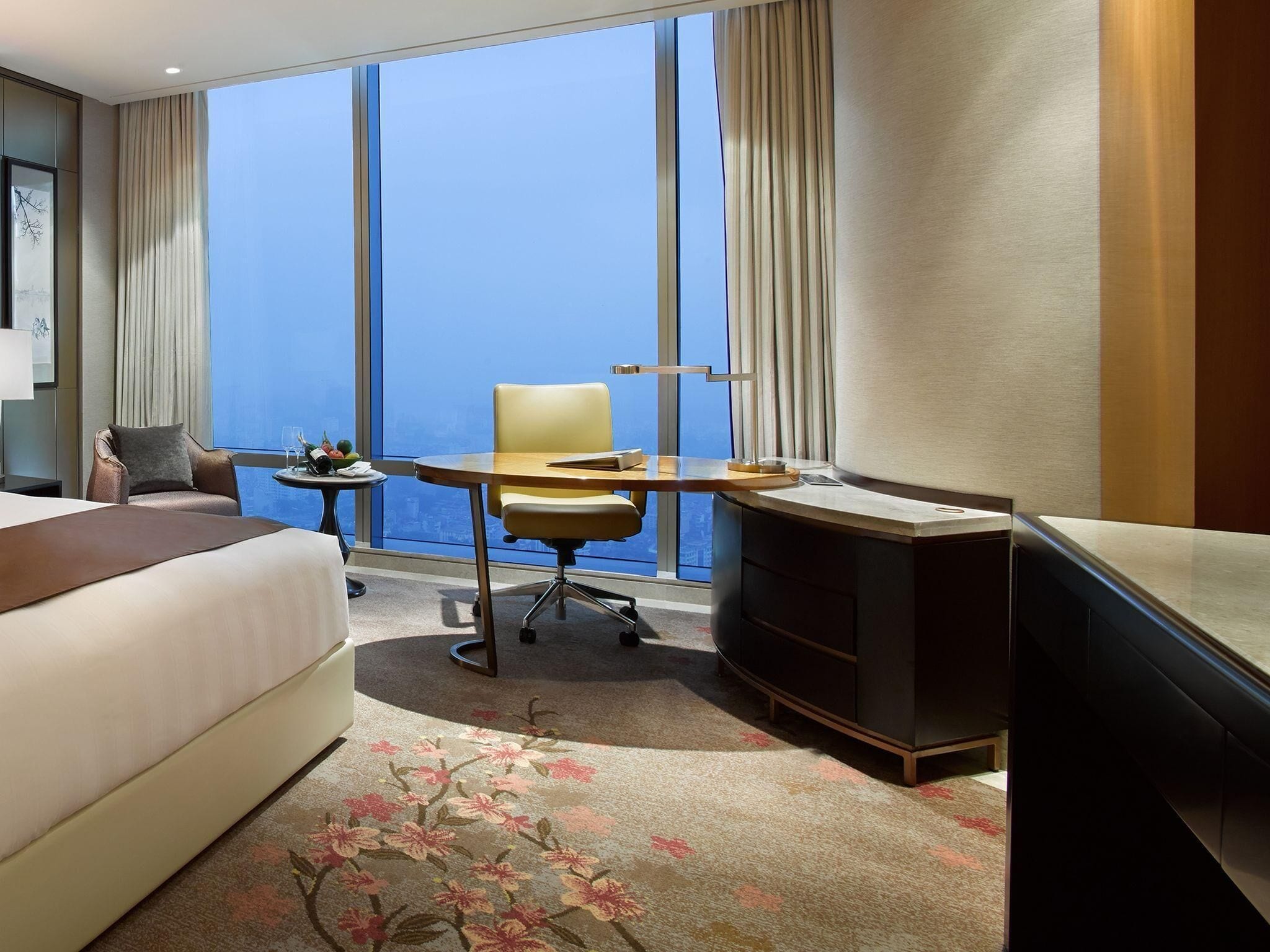A hotel room with a bed, a desk, a chair, a small table with a fruit bowl, and a view of the city through large windows.