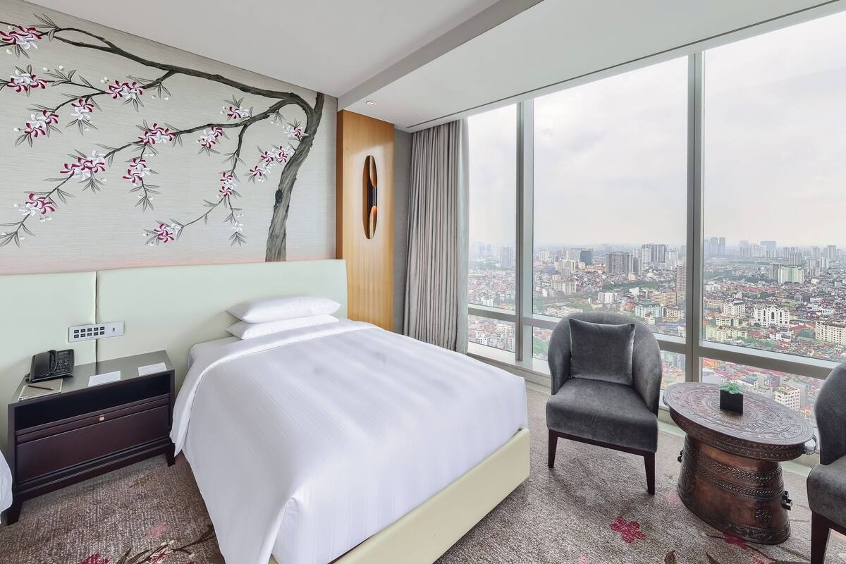 A hotel room with a large bed, a sitting area with a chair and a small table, a painting of a tree on the wall, and a city view through large windows.