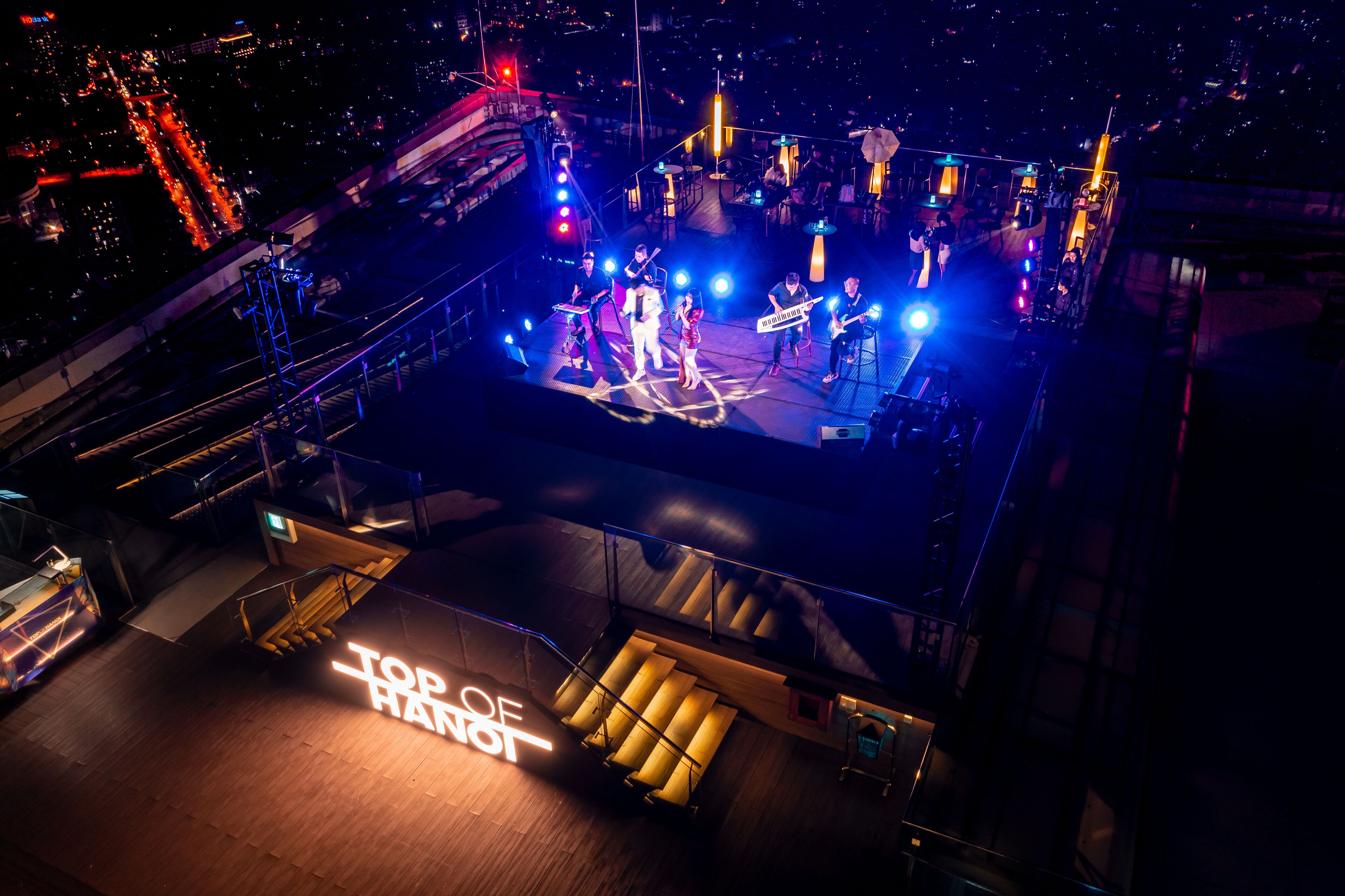 Un escenario en la azotea con una banda tocando, rodeado de asientos al aire libre e iluminado por luces de escenario.