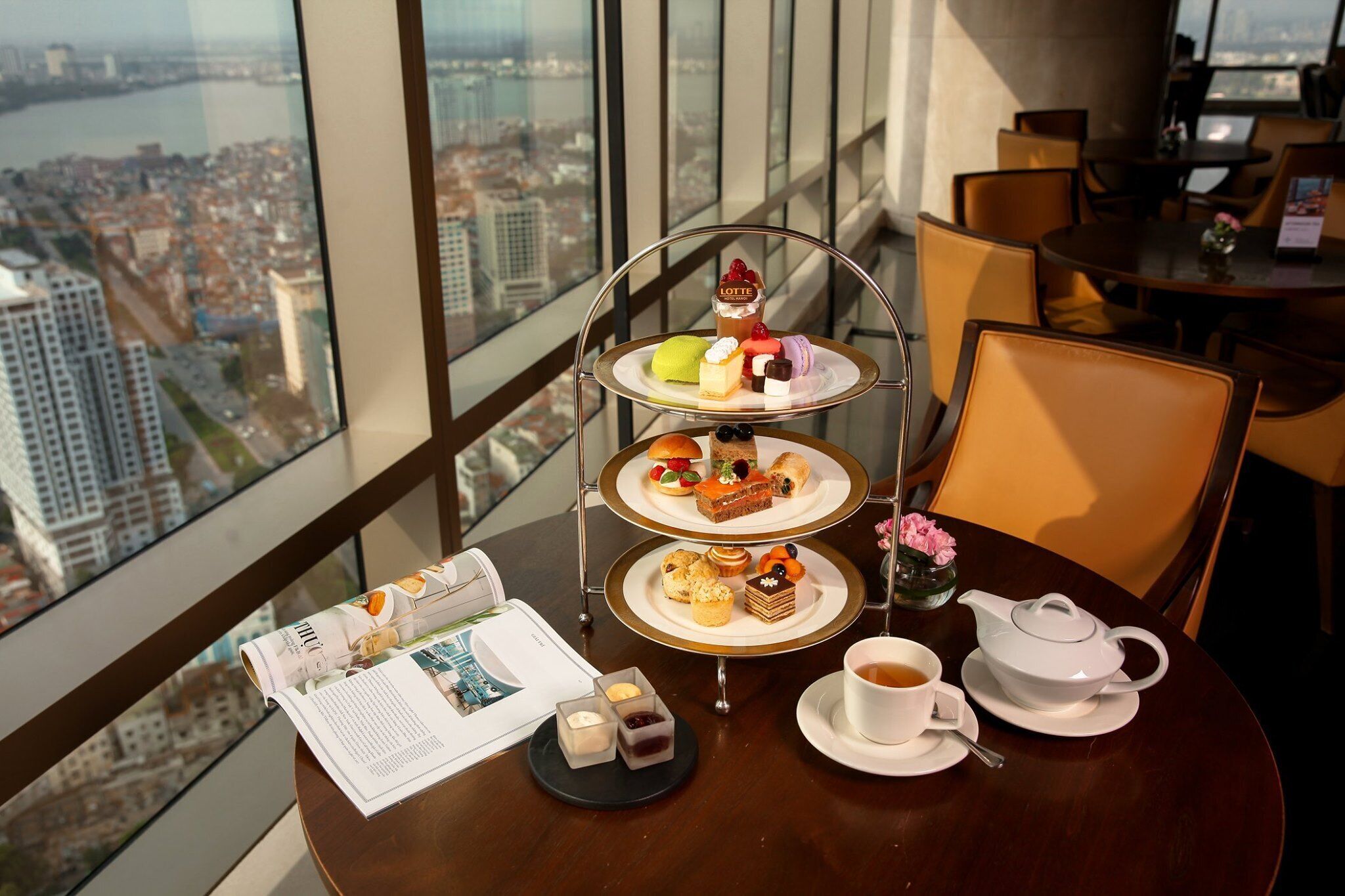 A three-tiered dessert stand with various pastries and a teapot on a table, with a city view through large windows.