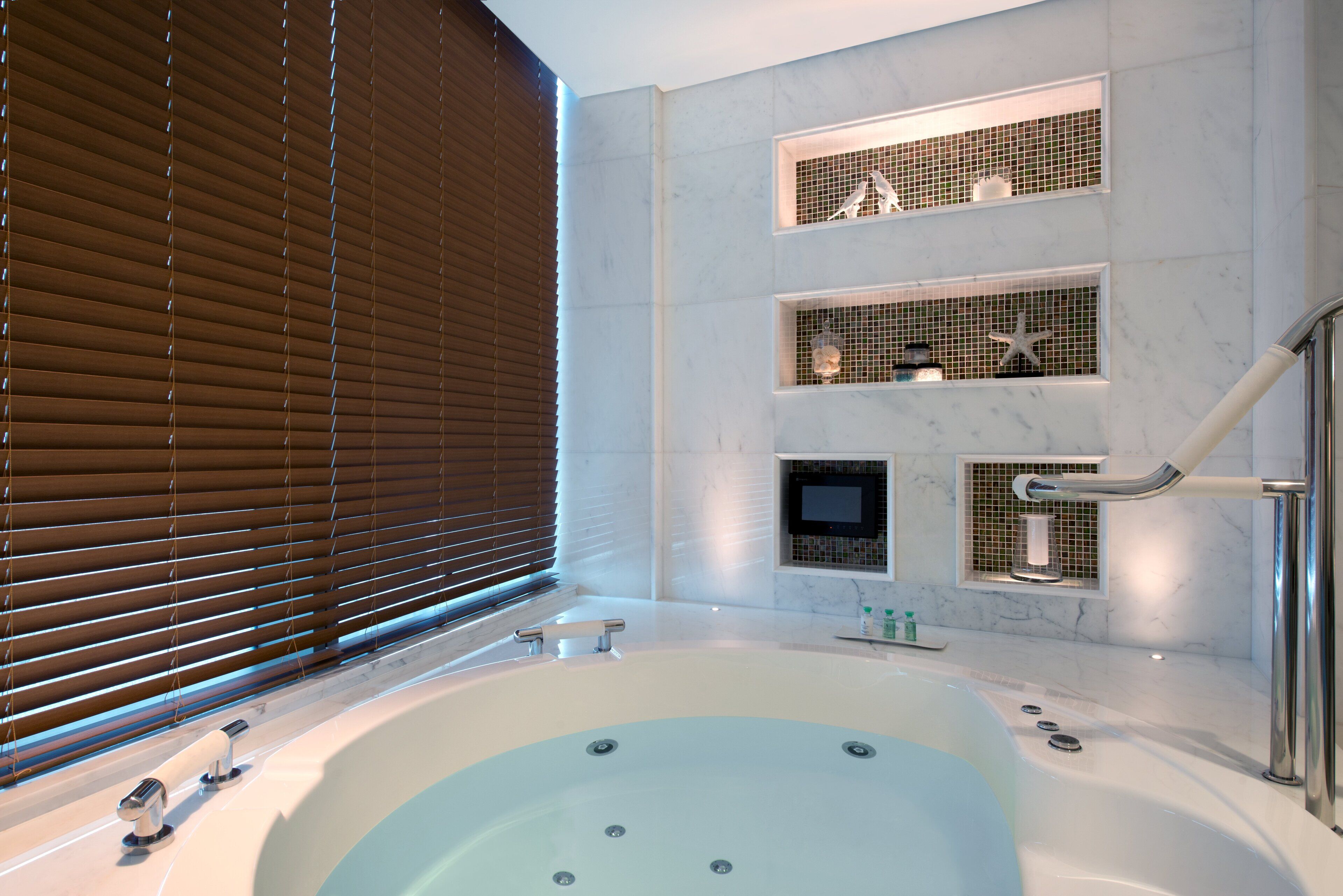 Un baño moderno con una bañera grande, estantes empotrados con objetos decorativos y una ventana con persianas.