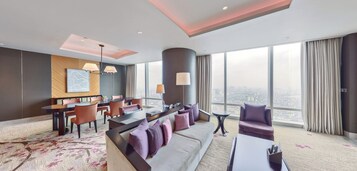 A modern hotel room with a large window offering a city view, a dining area with a table and chairs, and a seating area with a sofa and armchairs.