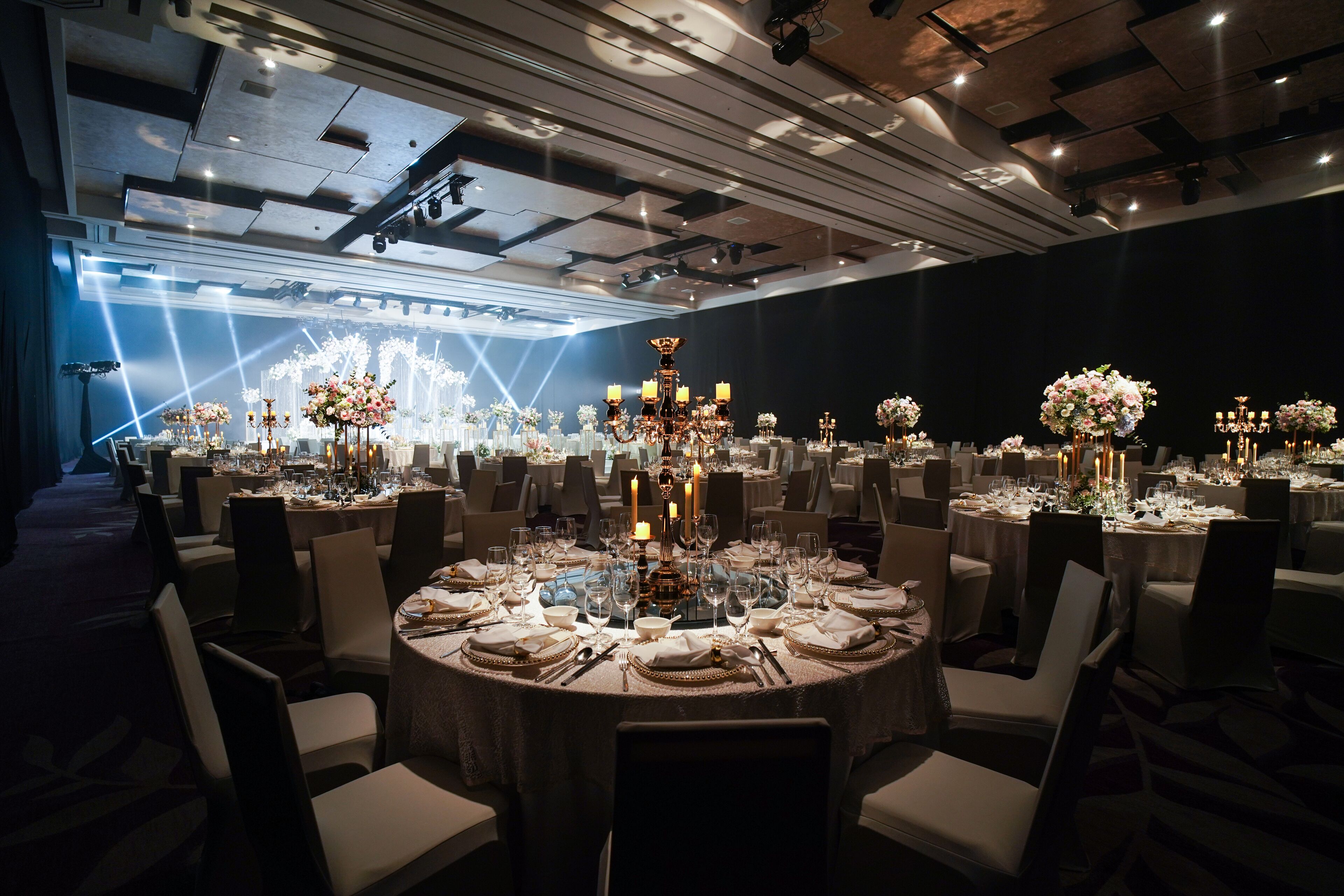 A banquet hall with round tables set for a formal event, featuring chandeliers and floral centerpieces.