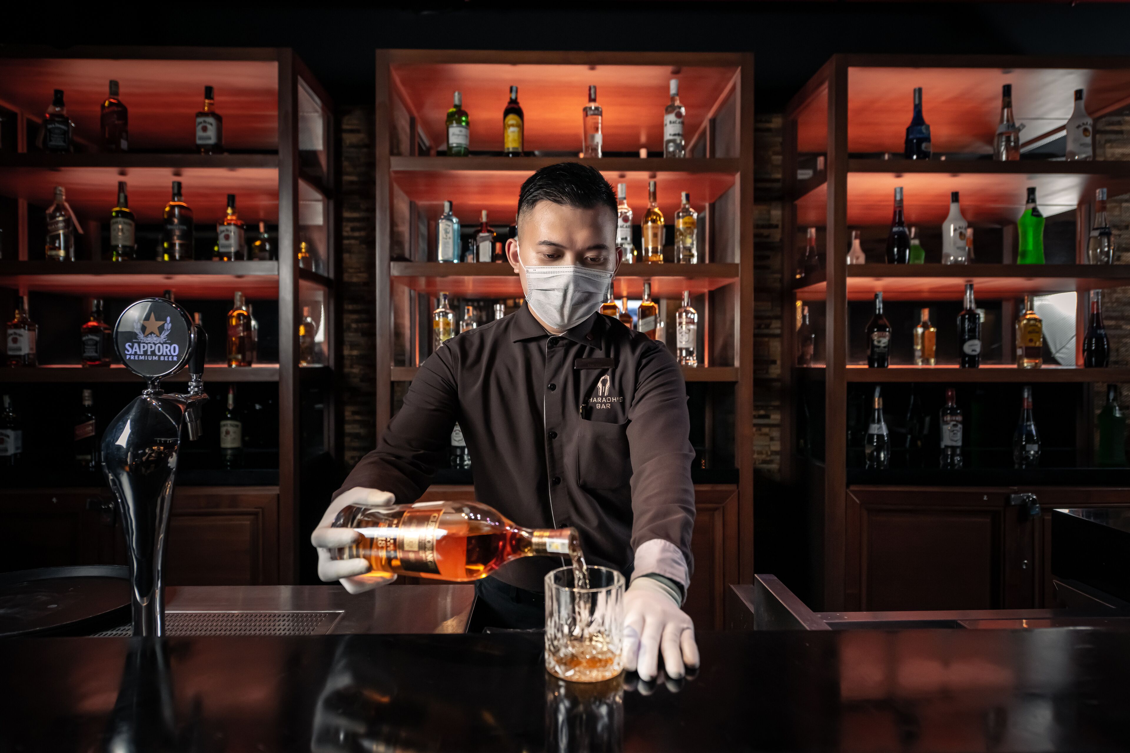 A bartender pouring a drink from a bottle into a glass at a bar with shelves of liquor bottles in the background.