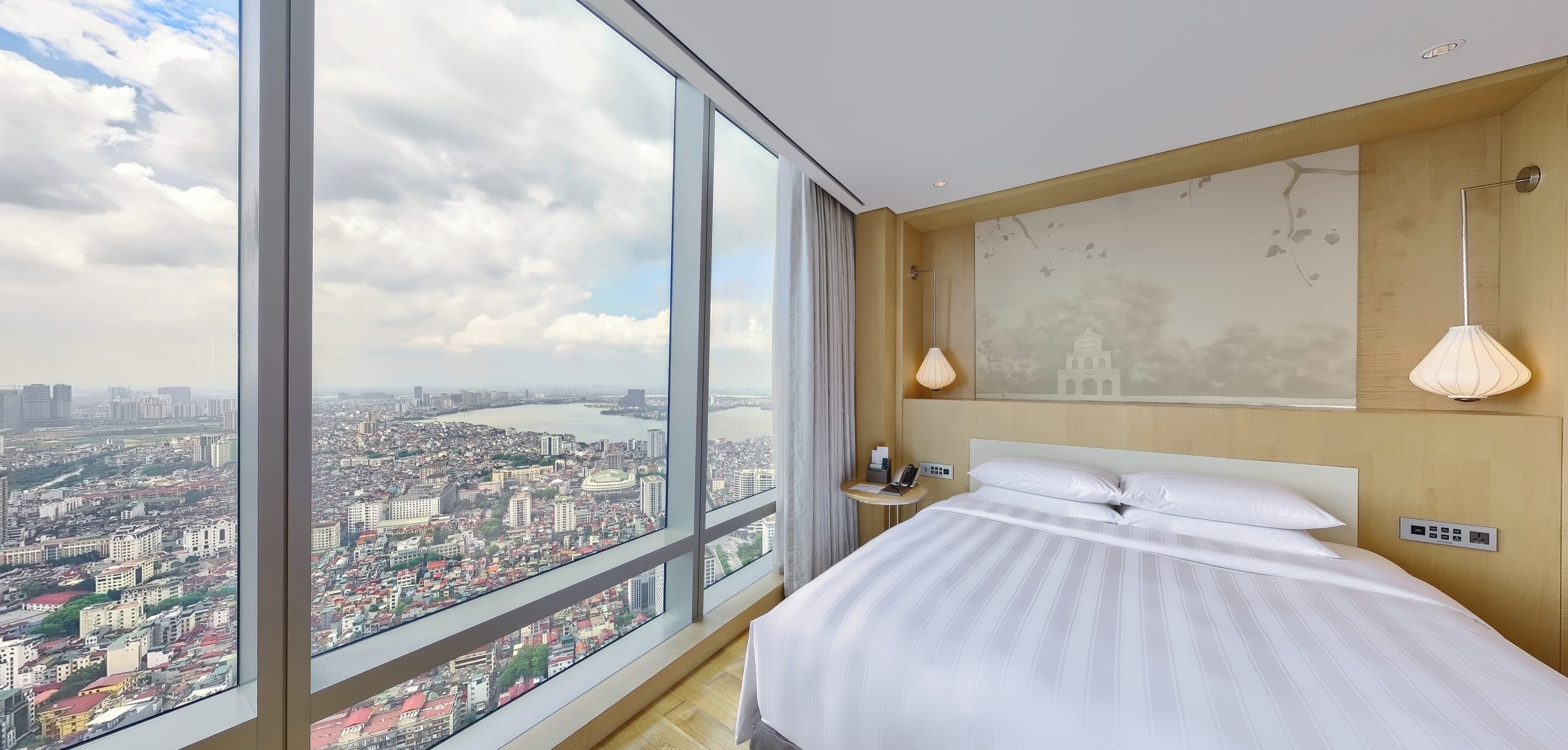 Habitación de hotel con un ventanal grande que ofrece vistas a la ciudad, una cama, mesita de noche, lámpara y teléfono montado en la pared.
