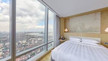 Habitación de hotel con un ventanal grande que ofrece vistas a la ciudad, una cama, mesita de noche, lámpara y teléfono montado en la pared.
