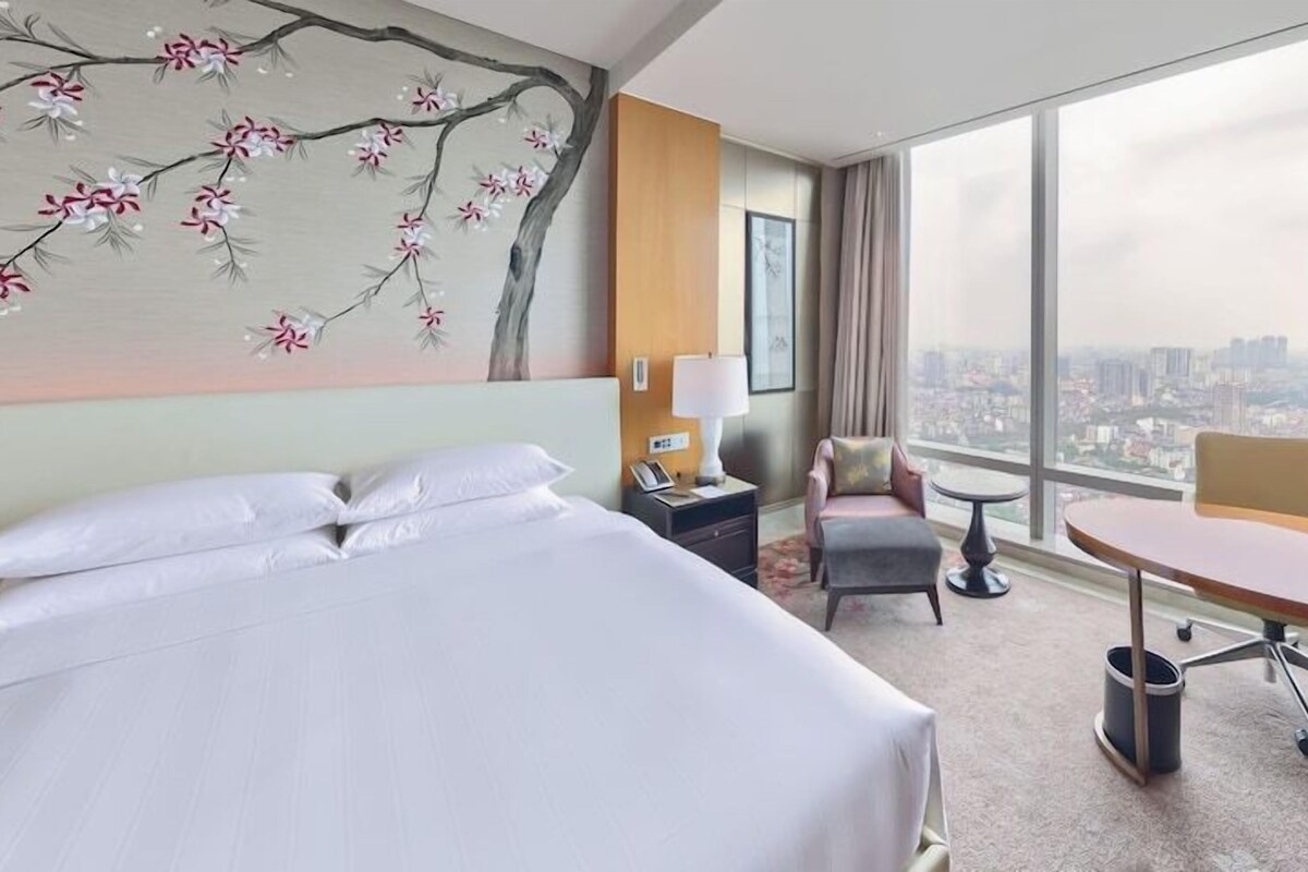 A hotel room with a large bed, a desk, a chair, a nightstand, and a view of the city.