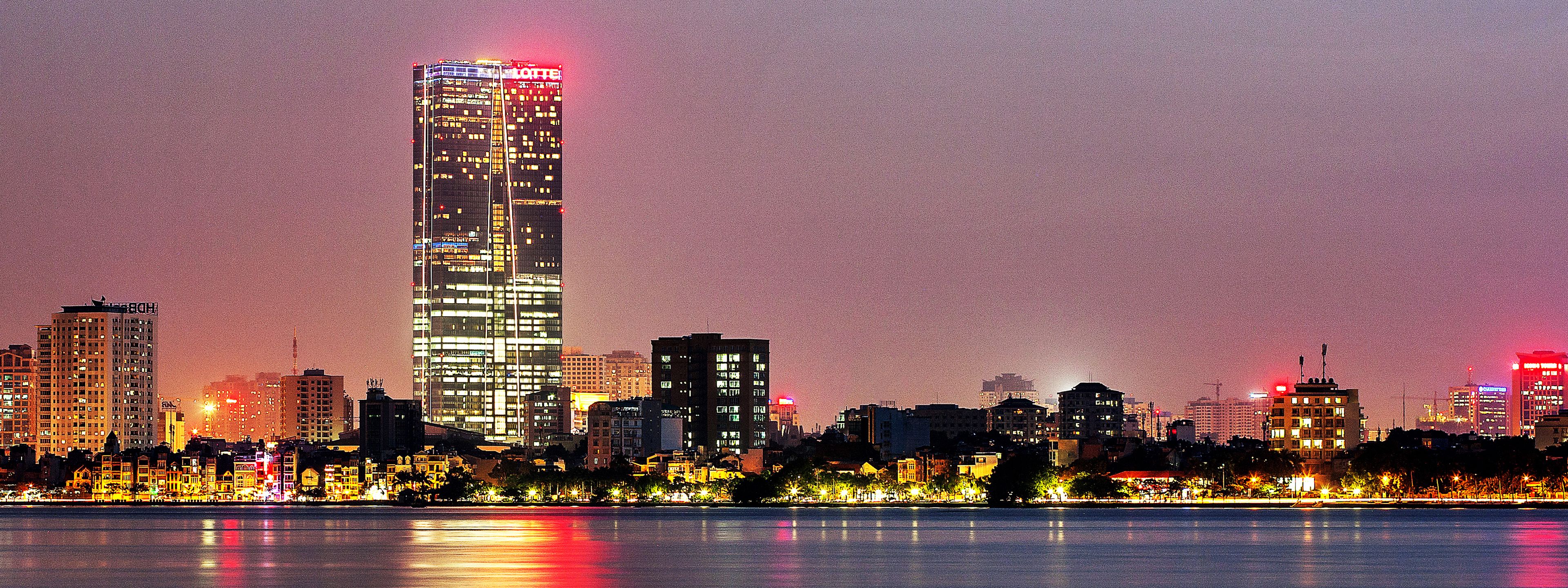 Un horizonte urbano con edificios iluminados, una masa de agua al frente y un cielo despejado.