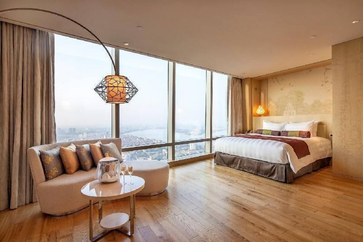 A modern hotel room with a large window, a sofa, a round coffee table, and a bed with a view of the city.