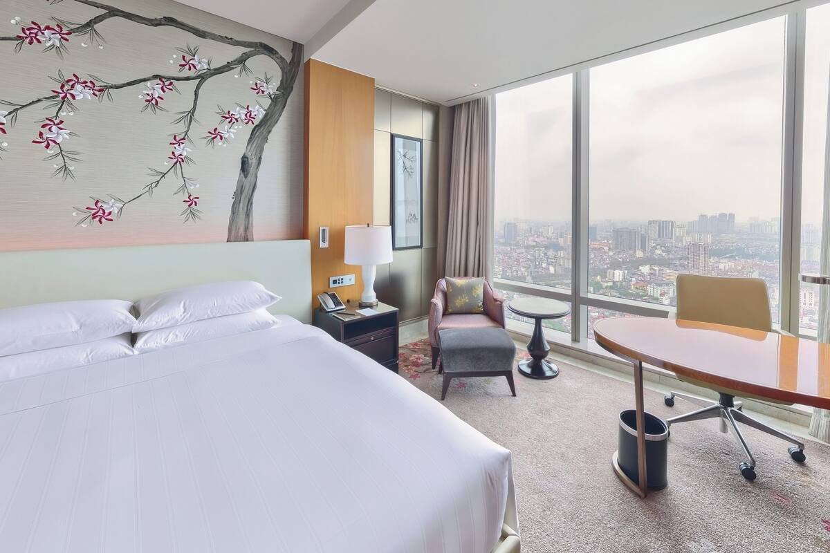 A hotel room with a large bed, a desk, a chair, a small table, and a view of the city through large windows.