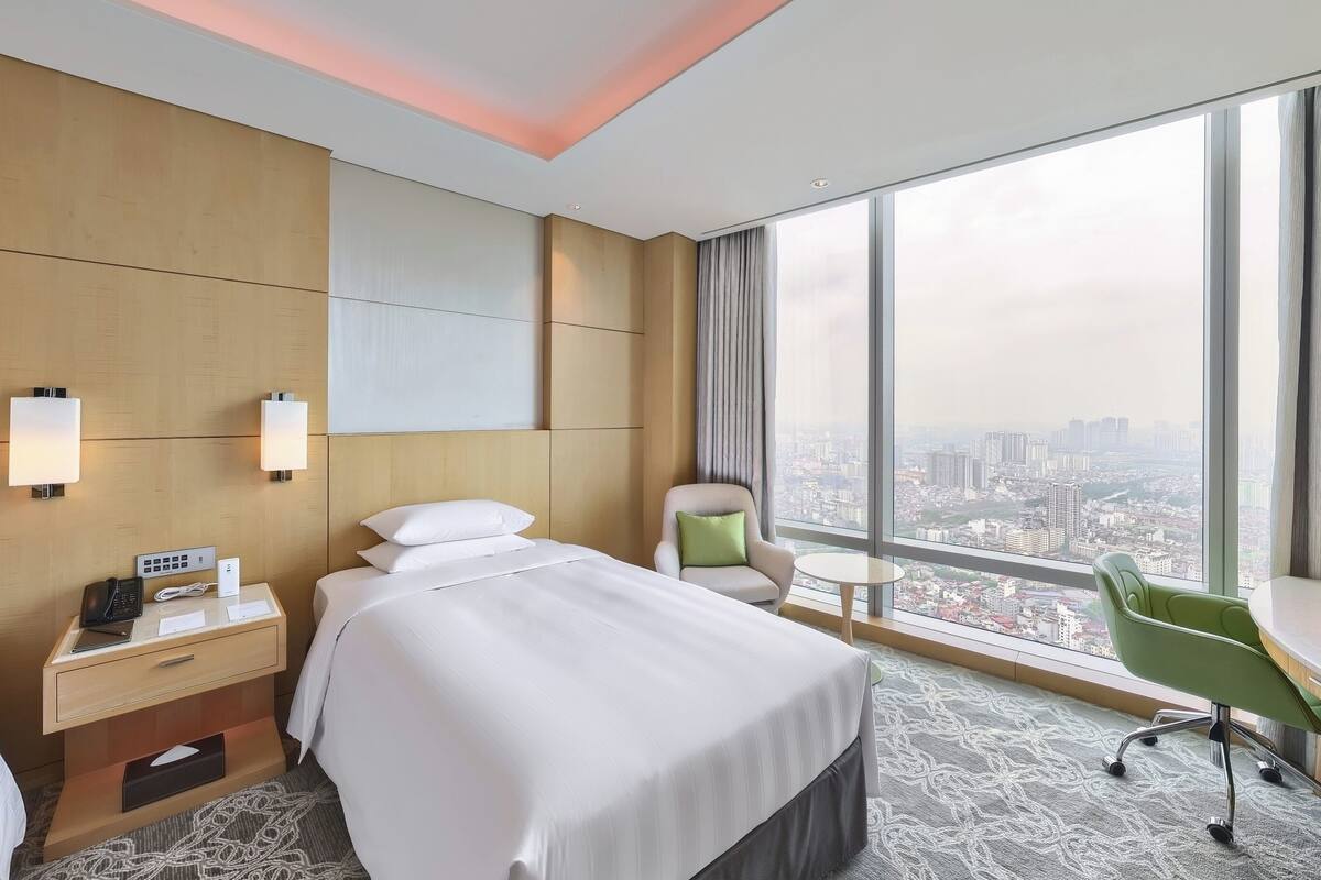 A hotel room with a bed, bedside table with a phone and lamp, a desk with a chair, and a large window offering a city view.