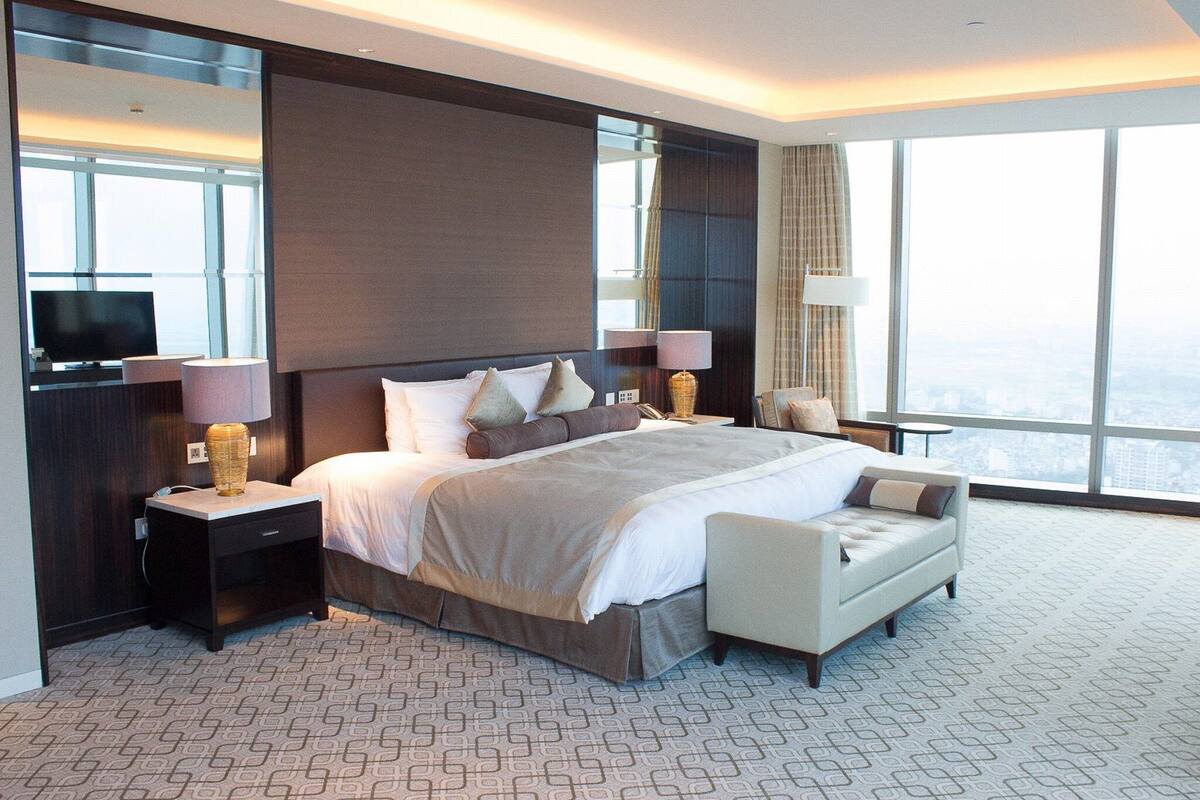 A modern hotel room with a large bed, two bedside tables with lamps, a small sofa, and a large window offering a city view.