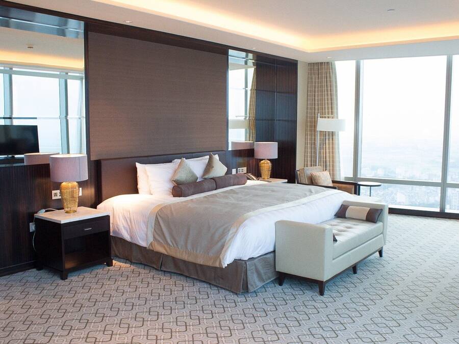 Una habitación de hotel moderna con una cama grande, dos mesitas de noche con lámparas, un pequeño sofá y un ventanal con vistas a la ciudad.