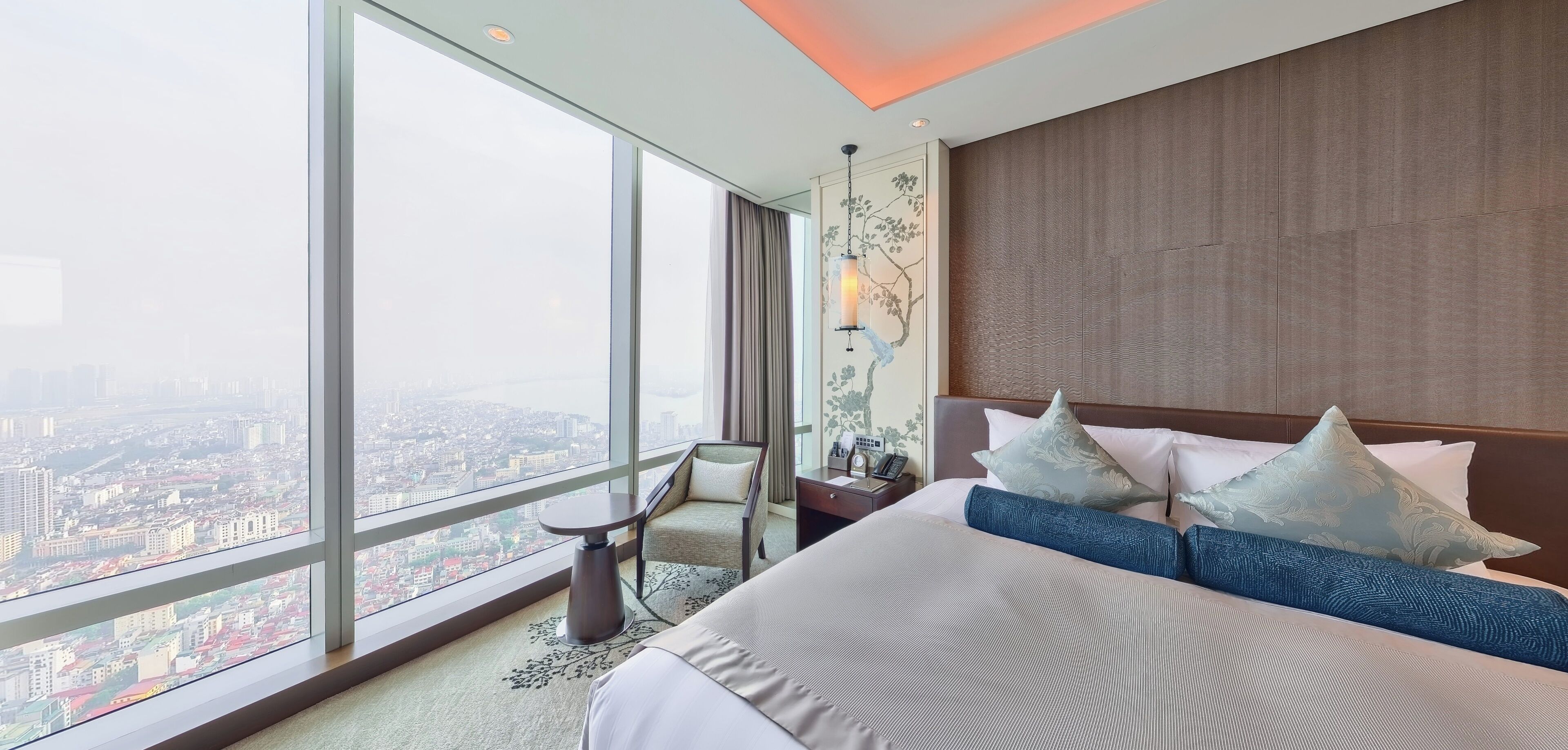 A hotel room with a large window offering a city view, a bed with blue and white bedding, a small table, and a chair.