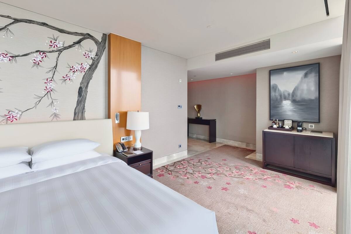 A hotel room with a bed, a nightstand with a lamp, a painting of a tree, a fireplace, and a television.