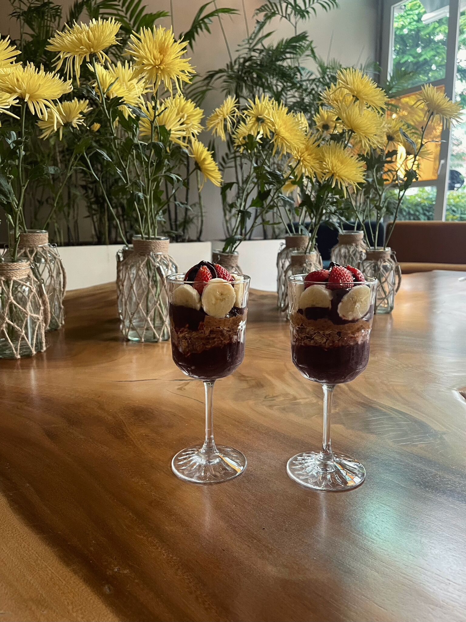 Dos vasos de postre sobre una mesa de madera con flores amarillas al fondo.