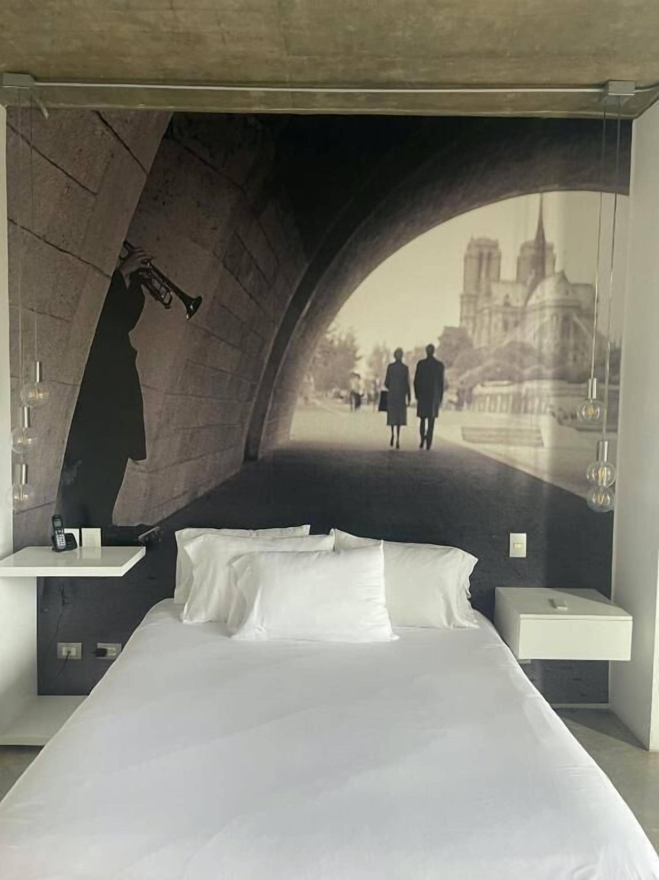 Una habitación de hotel moderna con una cama centrada debajo de un amplio ventanal en arco. En la pared detrás de la cama se ve un cartel con una vista de la ciudad y gente caminando.