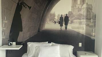Una habitación de hotel moderna con una cama centrada debajo de un amplio ventanal en arco. En la pared detrás de la cama se ve un cartel con una vista de la ciudad y gente caminando.
