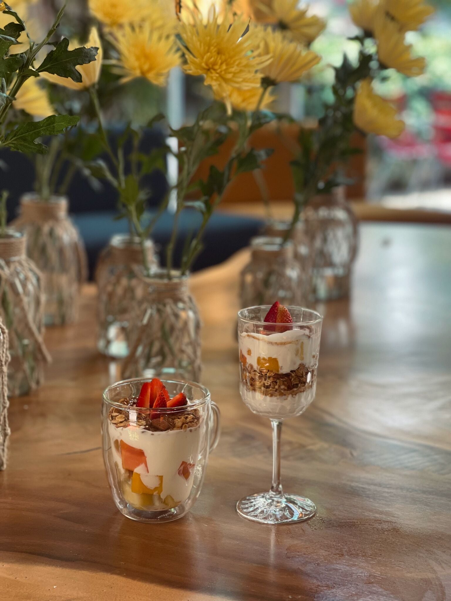 Una mesa con dos vasos de postre, uno con un postre en capas y el otro con fruta encima, frente a un fondo de flores amarillas en frascos de vidrio.