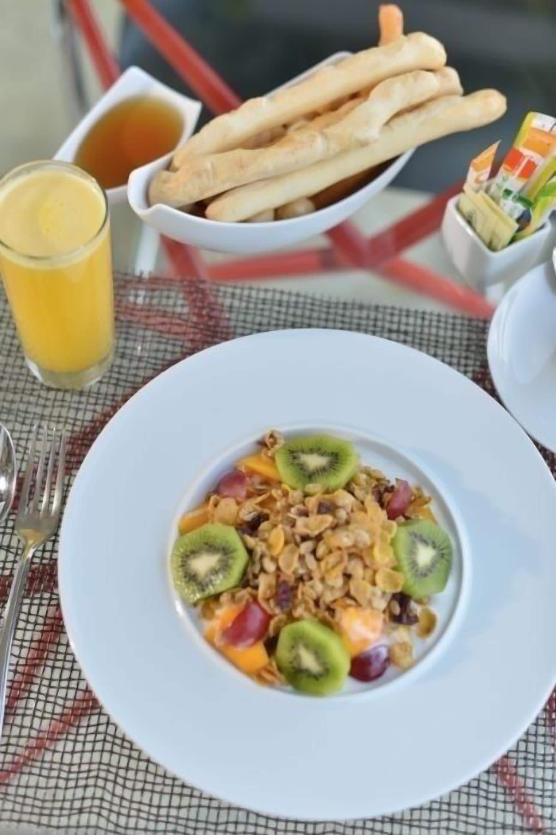 A breakfast setting with a plate of cereal topped with fruits, a glass of orange juice, and a bowl of breadsticks.