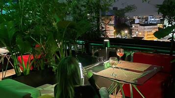 A person sitting on a green sofa at a table with a glass of wine, overlooking a cityscape at night.