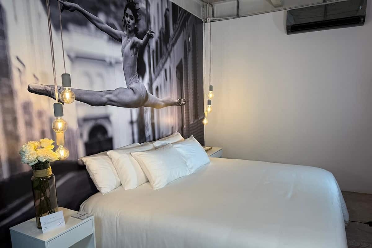 Una habitación de hotel moderna con una cama grande, ropa de cama blanca, una mesita de noche con un florero y un aplique de pared.