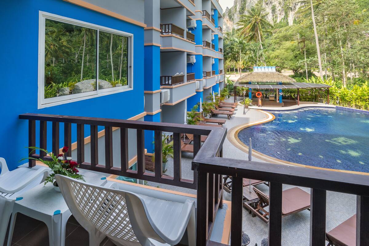 A hotel balcony with a pool, lounge chairs, and a view of the surrounding greenery.