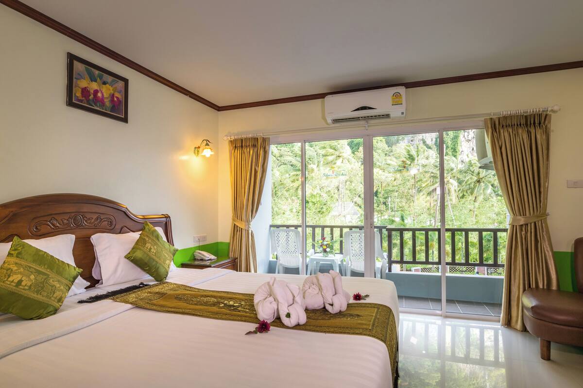 A hotel room with a large bed, a balcony with a view of greenery, a painting on the wall, and an air conditioning unit.