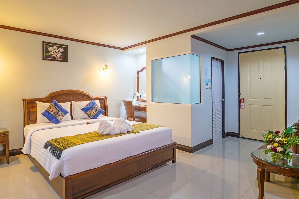 A hotel room with a wooden bed, bedside tables, a desk, a chair, a mirror, a wall-mounted TV, and a door.