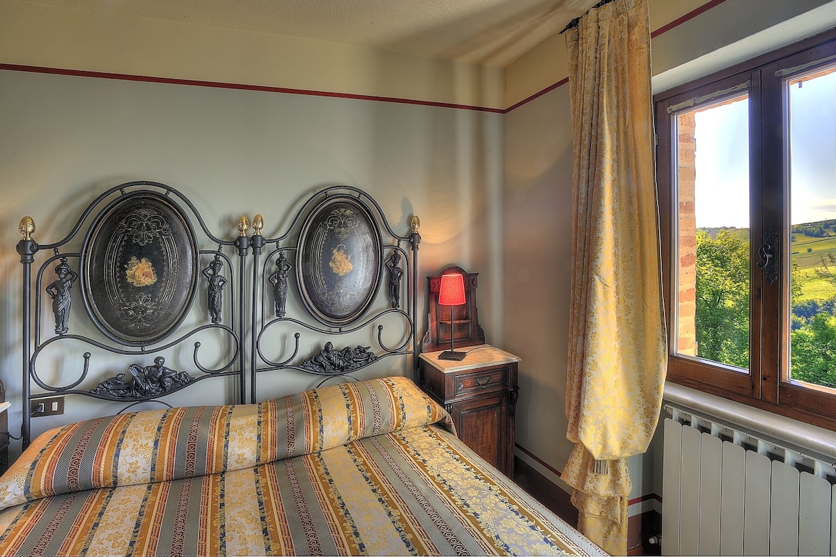Ein Schlafzimmer mit Bett, Nachttisch, Lampe und einem Fenster mit Blick auf Grünflächen.