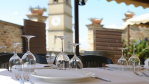 A table set for dining with wine glasses and cutlery, a clock tower in the background, and a brick wall. - Albergo Diffuso Borgo Montemaggiore (Colli al Metauro)