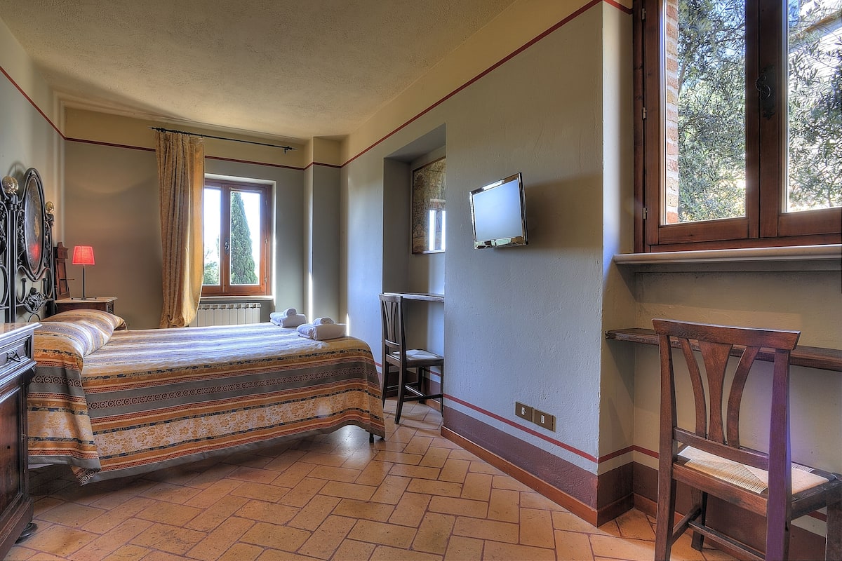 Ein Schlafzimmer mit Bett, Sessel, Fernseher und Fenster mit Blick auf Bäume.