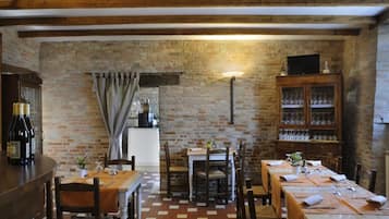 A rustic dining area with brick walls, wooden beams, and a tiled floor. There are tables set for dining, a bar area with bottles, and a cabinet with glassware.
