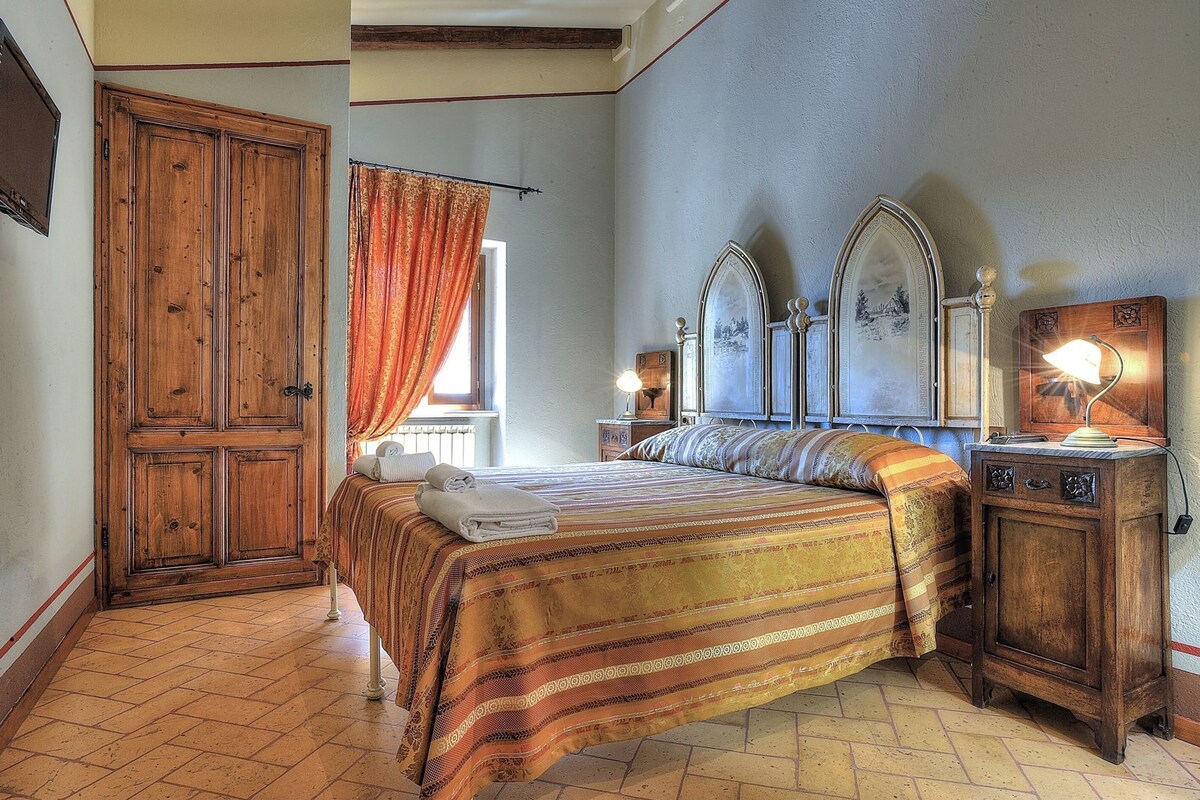 A bedroom with a large wooden wardrobe, a bed with a striped bedspread, two bedside tables with lamps, and a window with curtains.