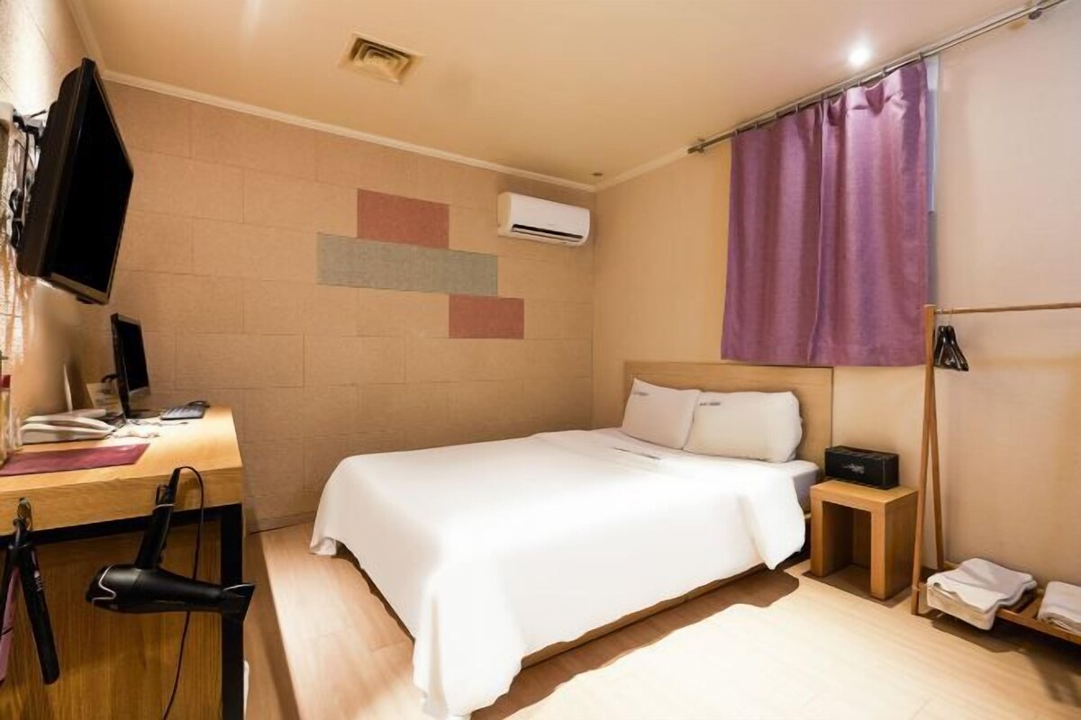 A hotel room with a bed, a desk, a chair, an air conditioning unit, and a television.