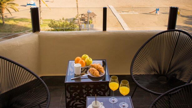 Ein Balkon mit Frühstückstisch, der Blick auf einen Strand mit Palmen und das Meer gewährt.