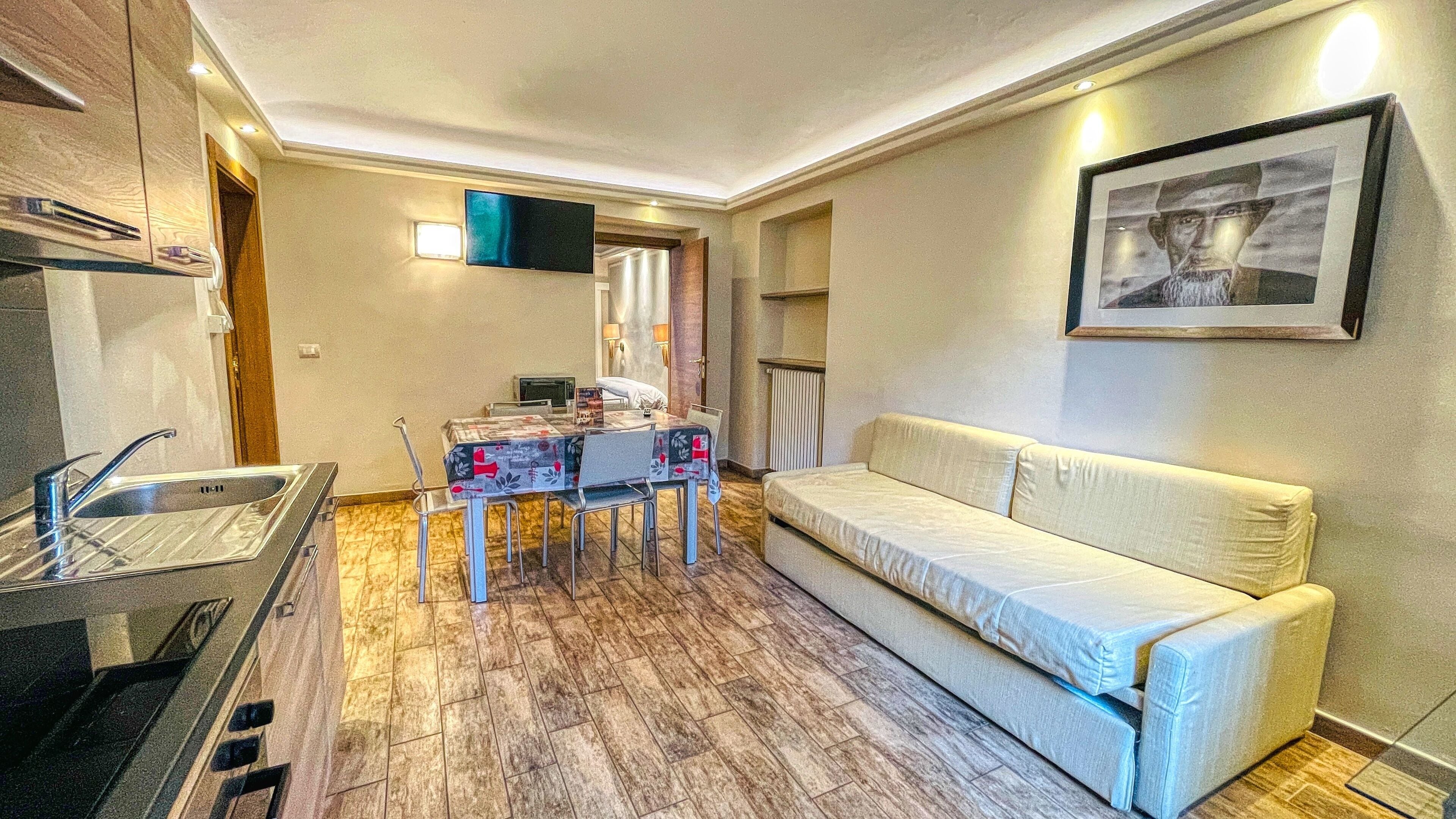 A compact living space with a kitchenette, a dining table, a sofa, and a flat-screen TV mounted on the wall.
