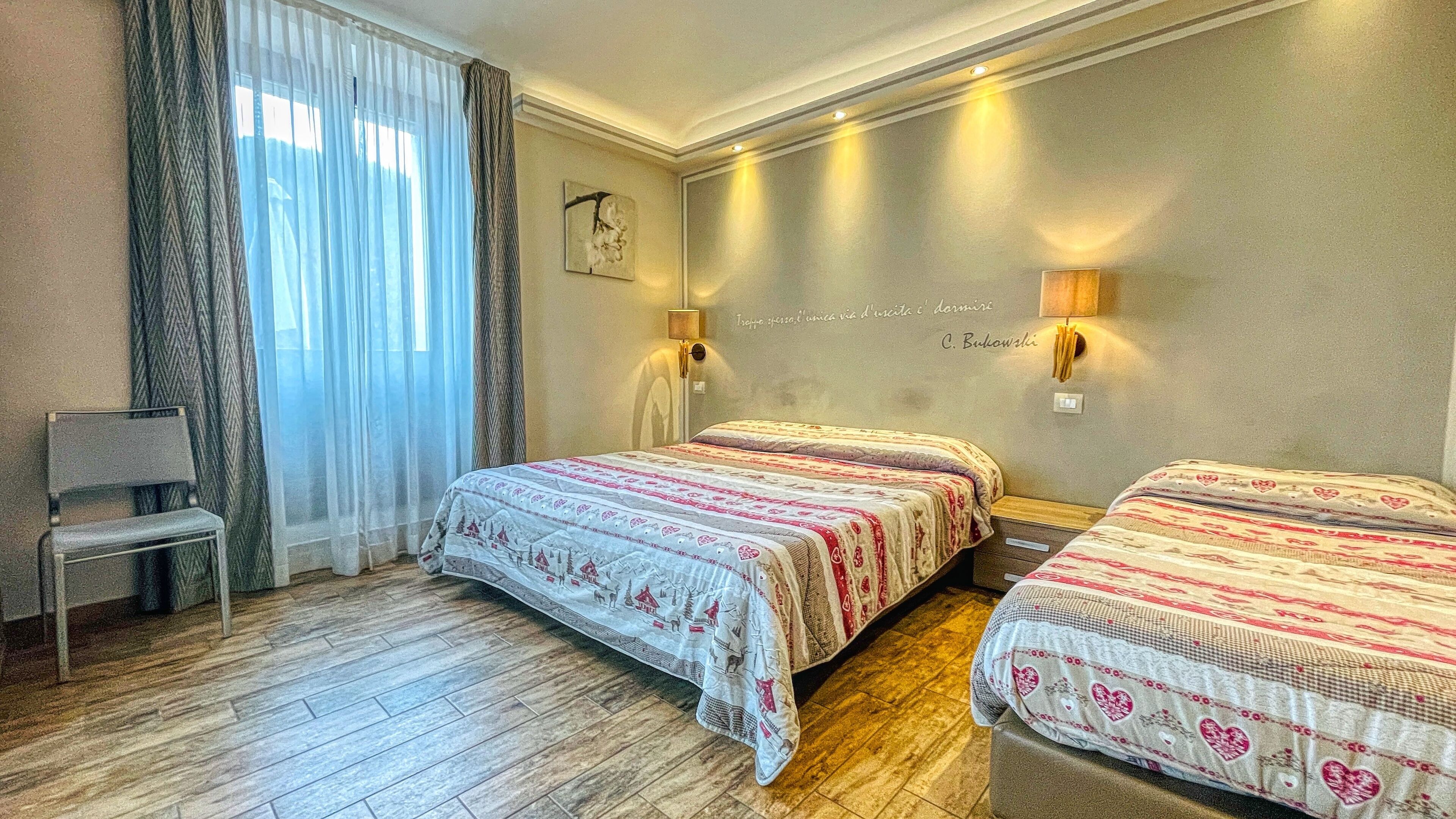 A hotel room with two beds, a chair, a large window with curtains, and a wall-mounted lamp.