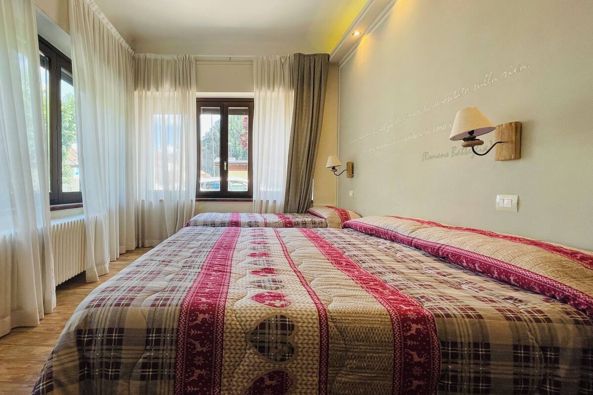 A hotel room with two beds, a window with curtains, a wall-mounted lamp, and a view of the outdoors.