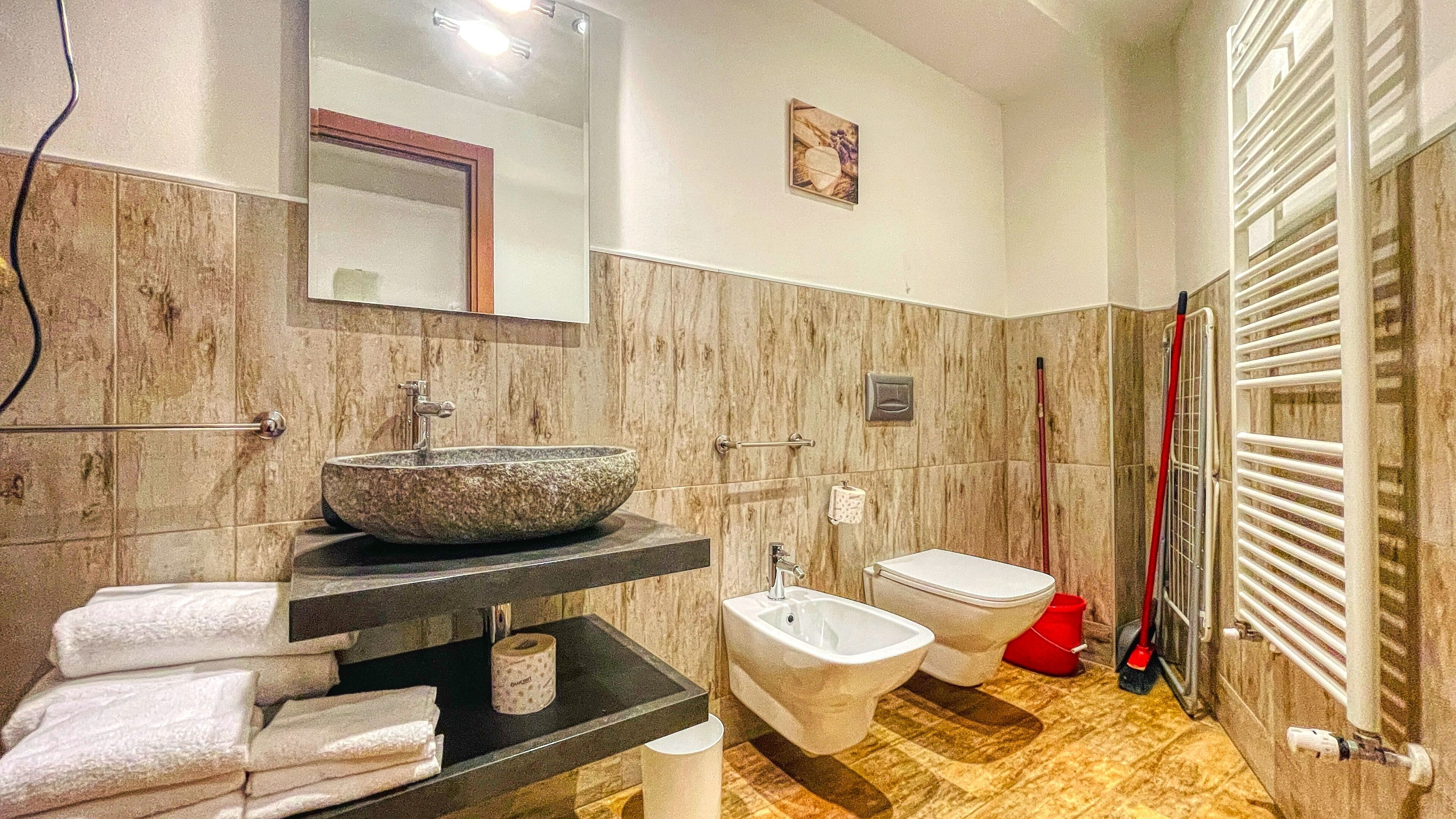A bathroom with a large stone sink, a bidet, a toilet, and a shower area with a red broom.