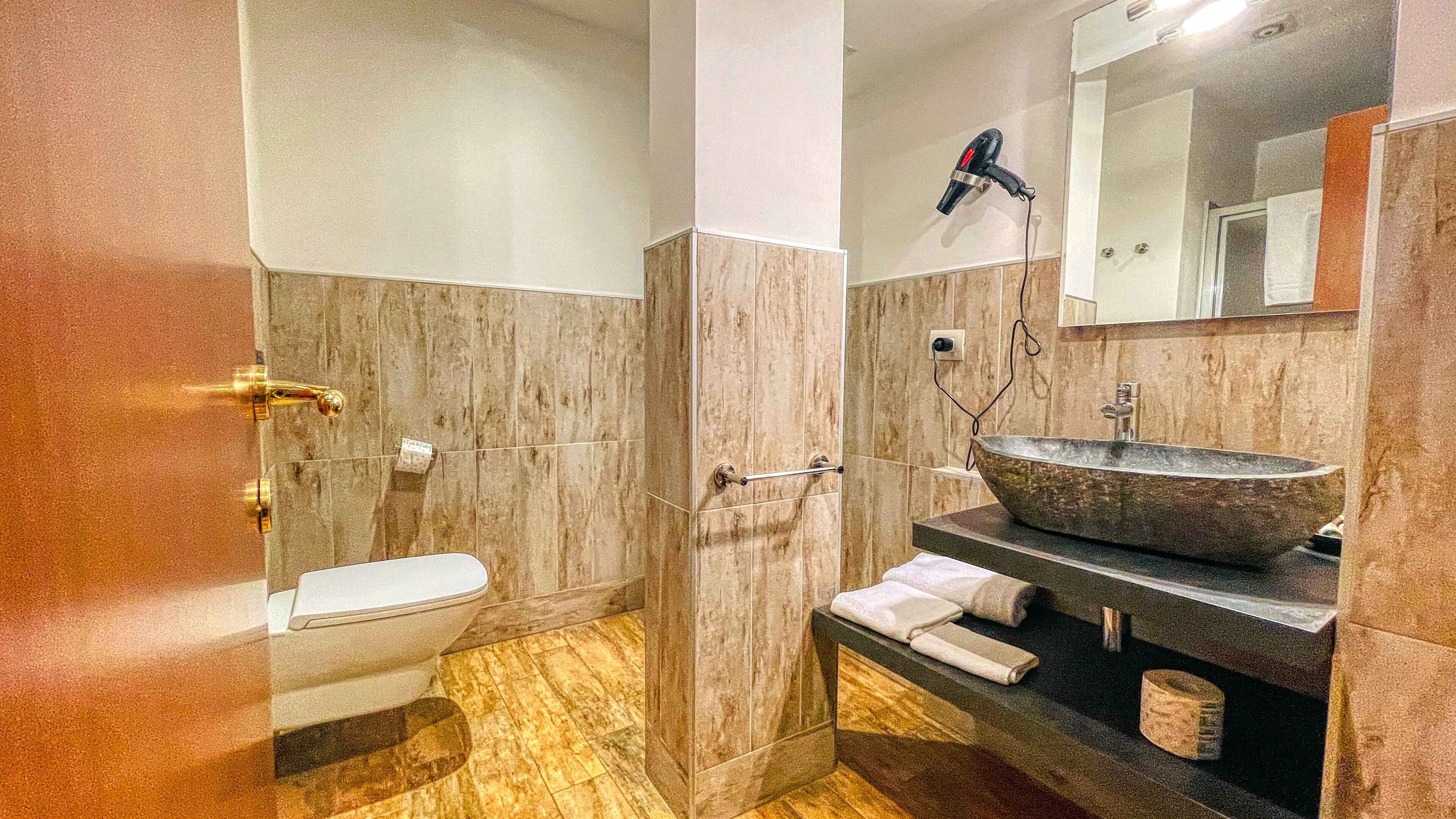 Une salle de bain avec un plancher en bois, des toilettes blanches, un grand miroir, un sèche-cheveux et un lavabo en pierre.