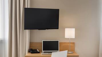 A hotel room with a desk, a laptop, a telephone, a television mounted on the wall, and a bedside lamp.