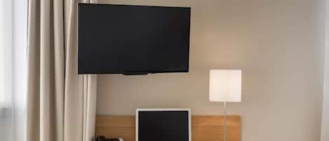 A hotel room with a desk, a laptop, a telephone, a television mounted on the wall, and a bedside lamp.