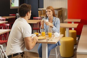 Ein Café mit Holztischen und roten Stühlen, einem gelben Ottomanen sowie einer roten Wand.