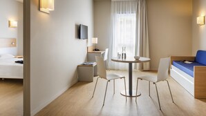A hotel room with a bed, a desk with a chair, a round table with chairs, a TV mounted on the wall, and a window with curtains. - Aparthotel Adagio Access Dijon République (Dijon)